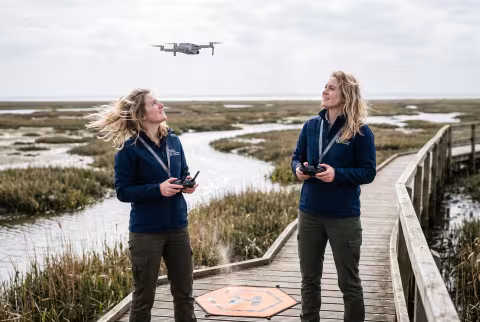 Research assistant launching a survey drone over a saltmarsh — Person `main_image`, student/researcher.